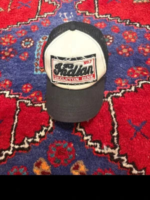 Svart keps  - Säljer en svart och vit truckerkeps från Skeleton Dink med broderad patch framtill där det står 'The Indian No.7'. Kepsen har mesh på sidorna och bak, samt böjd skärm. Perfekt för dig som gillar streetwear och retrostil.