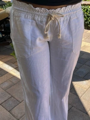 Vita linnebyxor från Roxy - Säljer ett par vita linnebyxor från Roxy med avslappnad passform, bred resår i midjan och snörning framtill. Byxorna har raka ben och stora fickor fram. Perfekta för en chill och stilren look.