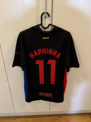 FC Barcelona Raphinha matchtröja svart - Snygg svart FC Barcelona matchtröja med Raphinha och nummer 11 på ryggen. Tröjan har klubbmärke och Spotify-logga framtill samt röda och blå detaljer på sidorna. Perfekt för fotbollsfans som vill sticka ut på läktaren eller planen.