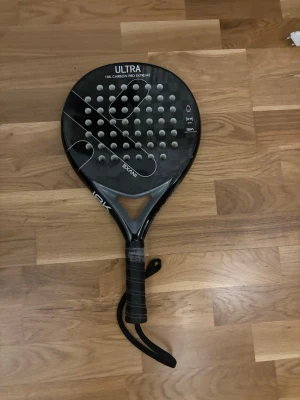 Wilson Ultra padelracket svart kolfiber - Padelracket från Wilson, modell Ultra 18K Carbon Pro Extreme. Racketen är svart med rund form, perforerad träffyta och handledsrem. Tillverkat i 18K kolfiber för extra styrka och kontroll. Perfekt för dig som vill ta padelspelet till nästa nivå.