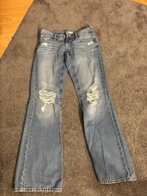 Ljusblå slitna bootcut jeans från Gina 14+ - Säljer ett par ljusblå bootcut jeans med slitningar och hål på båda knäna. Jeansen har klassisk femficksdesign och är gjorda i denim. Perfekta för en avslappnad och trendig look. Endast provade