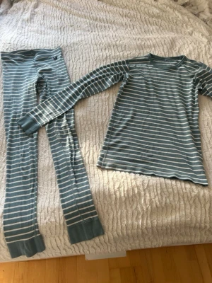 Randig grön pyjamas i bomull - Mysig tvådelad pyjamas med långärmad tröja och byxor i grön med vita ränder. Tillverkad i mjuk bomull, perfekt för chillkvällar eller sovmorgnar. Klassisk passform och bekväma muddar vid ärmar och benslut.