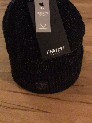 Svart stickad mössa från Lindberg - Snygg svart stickad mössa från Lindberg med diskreta reflexdetaljer. Klassisk ribbstickad design som passar perfekt till kalla dagar. Märket är broderat framtill och materialet känns mjukt och värmande.