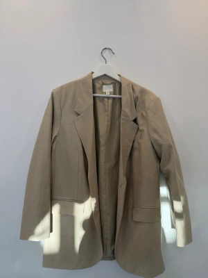 Beige kavaj från H&M - Stilren beige kavaj från H&M med klassisk krage och två stora fickor framtill. Rak passform och lätt material som ger en avslappnad men ändå dressad look. Perfekt att slänga över en t-shirt eller skjorta.