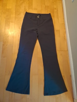 Mörk grå utsvängda kostymbyxor - Snygga svarta kostymbyxor med utsvängda ben och hög midja. Byxorna har två bakfickor med lock och knapp, samt klassisk knapp- och dragkedjestängning fram. Perfekta för en stilren look. Materialet känns mjukt och följsamt.