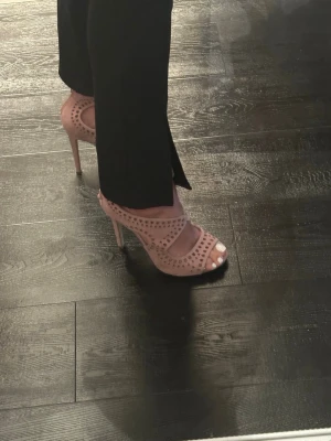 Rosa klackade sandaler från JustFab - Säljer ett par rosa sandaler från JustFab med hög klack och öppen tå. Skorna har snygga utskurna detaljer, snörning framtill och dragkedja bak. Materialet är textil med mockakänsla och de har en feminin, trendig look. Oanvända endast provade 