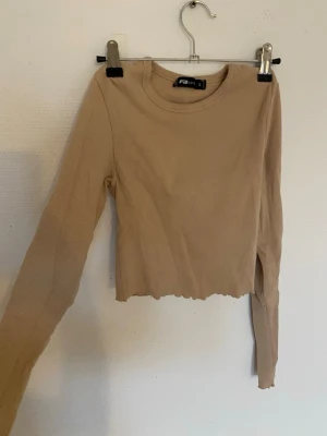 Beige ribbad croppad topp Junkyard - Trendig beige croppad topp från Junkyard med lång ärm och ribbad struktur. Toppen har en vågig kant nertill som ger en cool detalj. Perfekt att styla med höga jeans eller kjol för en avslappnad look.