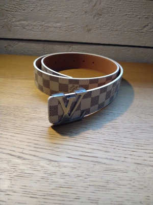 Rutig Louis Vuitton-bälte i vitt - Snyggt bälte från Louis Vuitton med klassiskt rutigt mönster i vitt och beige. Bältet har ett stort LV-spänne i silverfärg och är gjort i skinn. Perfekt accessoar för att lyfta din outfit.