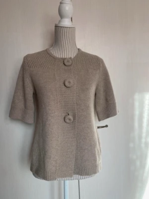 Beige stickad kofta med stora knappar - Beige stickad kofta med tre stora, runda knappar framtill. Koftan har korta ärmar, rund halsringning och ribbstickade detaljer vid ärmslut och nederkant. Mjuk och stilren design, perfekt för lager-på-lager.