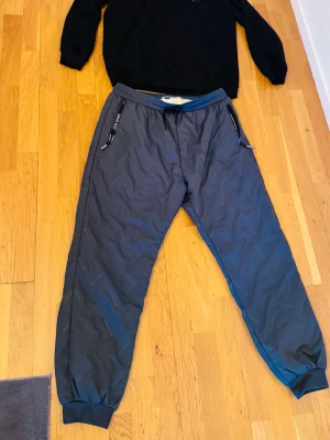 Fodrade täckbyxor och fleece tröja - Säljer ett par gråa täckbyxor med resår och dragsko i midjan samt muddar vid bensluten. Byxorna har två dragkedjeförsedda fickor med svarta detaljer och text på banden. Tillhörande svart fleecetröja    Bägge är fodrade. Perfekta för kalla dagar och outdoor-aktiviteter. Oanvända 