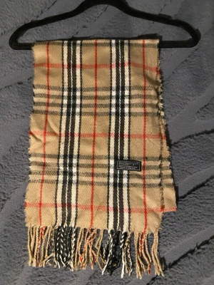 Klassisk Burberry Halsduk - Burberry halsduk 🤎  Klassisk Burberry-halsduk i det välkända rutmönstret. En tidlös favorit som aldrig går ur stil.  ✨ Detaljer: – Material: Lammull – Färg: Brun/Svart/Vit/Röd – Mått: 155 cm x 27 cm – Skick: Mycketbra – Original: ja  Perfekt till höst & vinter och funkar både casual och uppklätt. Skickas snabbt 📦  Pris kan diskuteras.