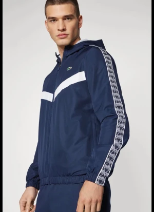 Marinblå vindjacka från Lacoste - Snygg marinblå vindjacka med vit diagonal detalj över bröstet och vita mönstrade ränder längs ärmarna. Jackan har huva, dragkedja och meshfoder på insidan. Klassisk Lacoste-logga på bröstet.