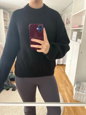 Svart stickad tröja från Lindex  - En svart stickad tröja med rund hals och långa ärmar. Tröjan har en enkel och stilren design, perfekt för lager-på-lager och passar till många olika outfits. Materialet ser mjukt och lite fluffigt ut.