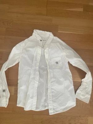 Vit skjorta från Diesel - Klassisk vit skjorta från Diesel med lång ärm och knappar framtill. Skjortan har en bröstficka med diskret Diesel-logga och är tillverkad i ett lätt bomullsmaterial. Perfekt för en stilren och enkel look.