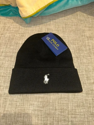 Polo Ralph Lauren - Svart ribbstickad mössa från Polo Ralph Lauren med klassisk uppvikt kant och broderad vit logga framtill. Perfekt accessoar för att hålla värmen och ge en stilren touch till din outfit.