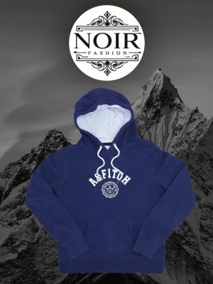 Mörkblå hoodie från A&Fitch - Snygg mörkblå hoodie från A&Fitch med vit logga och text på bröstet. Hoodien har en stor huva med vita dragsnören och en mjuk, ljus insida. Klassisk känguruficka framtill och ribbade muddar vid ärmar och nederkant. Perfekt för en avslappnad och trendig look.