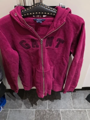 Vinröd hoodie med dragkedja från GANT - Säljer en vinröd hoodie från GANT med dragkedja framtill och stor huva. Hoodien har mjukt foder, fickor på sidorna och GANT-logga i stora bokstäver på bröstet. Perfekt för dig som vill ha en chill och snygg look.