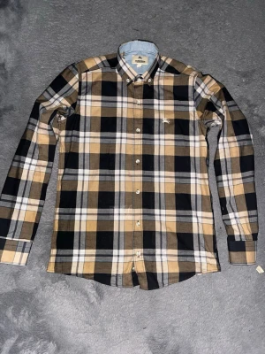 Rutig skjorta från Burberry - Säljer en klassisk rutig skjorta från Burberry i beige, svart och vit. Skjortan har långa ärmar, knappar framtill och en liten broderad logga på bröstet. Perfekt för dig som gillar stilrena och tidlösa plagg. Använd bara en gång!