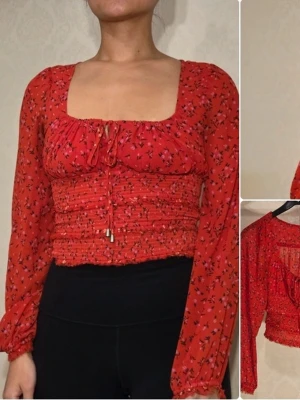 Röd blommig blus från Free People - Trendig röd blus med små svarta och vita blommor. Blusen har långärmade, transparenta ärmar och smockad midja. Söt detalj med knytning framtill och fyrkantig urringning. Perfekt för att lyfta vilken outfit som helst.