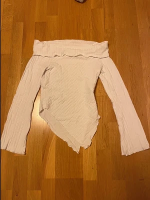 Vit offshoulder topp från AWARE - Trendig vit offshoulder topp från AWARE i ribbad struktur. Toppen har lång ärm och en unik asymmetrisk nederkant som ger en cool look. Perfekt för dig som vill sticka ut lite extra med din outfit.