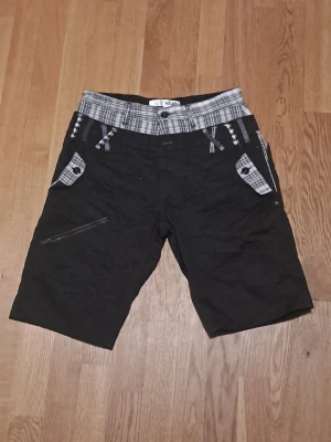 Svarta shorts med rutiga detaljer - Svarta shorts från Halham med gråsvart rutigt mönster i midjan och på fickorna. 