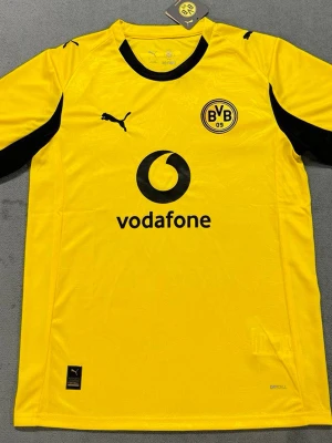 Gul Borussia Dortmund matchtröja Puma - Snygg gul Borussia Dortmund matchtröja från Puma med svart logga och detaljer. Tröjan har klubbmärke på bröstet, svart Vodafone-tryck och texten DORTMUND på ryggen. Tillverkad i lätt och ventilerande material, perfekt för fotboll eller träning.