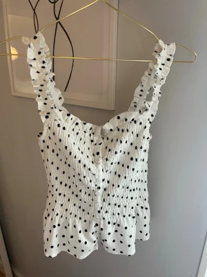 Vit prickig linnetopp med volang - Supersöt vit linnetopp med svarta prickar och volangdetaljer på axelbanden. Smockad överdel och knappar framtill ger en skön och stretchig passform. Perfekt för varma dagar och enkel att styla till jeans eller kjol.