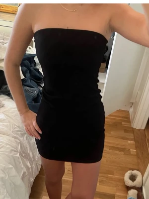 Svart axelbandslös kort klänning - Snygg svart axelbandslös klänning med tight passform och kort längd. Perfekt för dig som gillar en enkel men stilren look. Klänningen är ärmlös och har en rak, minimalistisk design som framhäver figuren.