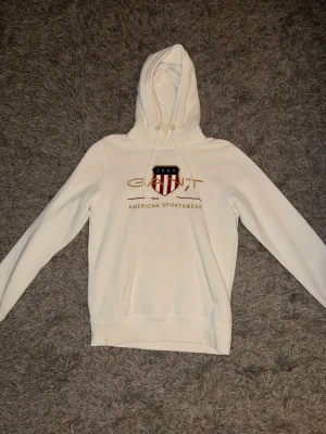 Vit hoodie från Gant - Snygg vit hoodie från Gant med stor broderad logga och sköld på bröstet. Storlek S, Klassisk känguruficka framtill, dragsko i huvan och ribbade muddar. Tillverkad i mjukt bomullsmaterial för en bekväm och avslappnad look. Perfekt för dig som gillar stilren streetwear. Ny pris 1200kr