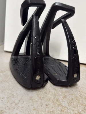 Svarta stigbyglar  - Säljer ett par svarta stigbyglar  i tålig plast. De har en greppvänlig yta med små piggar på fotplattan och modern design. Perfekta för dig som rider och vill ha lätta och funktionella stigbyglar.