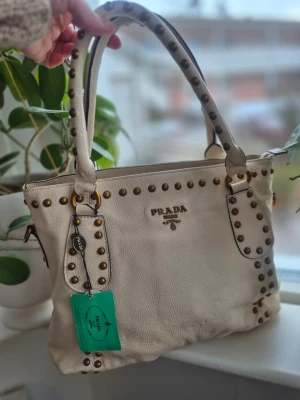 Vit handväska med nitar från Prada - Säljer en snygg cremefärgad handväska från Prada i äkta läder. Väskan har guldfärgade nitar längs kanterna och på handtagen, samt Pradas logga i guld framtill. Två rejäla handtag gör den enkel att bära. Perfekt för dig som gillar exklusiva accessoarer.