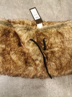Moccafärgad fuskpäls sjal från Pieces - Säljer en NY moccafärgad sjal i mjuk fuskpäls från Pieces. Sjalen har ett band för knytning och ger en lyxig känsla till din outfit. Perfekt för att hålla värmen och addera stil till vinterlooken. Ny me lappar, ordinarir pris 199kr