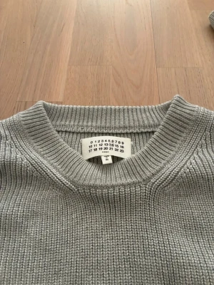 Grå stickad tröja från Maison Margiela - Säljer en stilren grå stickad tröja från Maison Margiela. Tröjan har ribbstickad struktur, rund halsringning och de ikoniska vita sömmarna i nacken. Perfekt för dig som gillar minimalistisk och exklusiv design.
