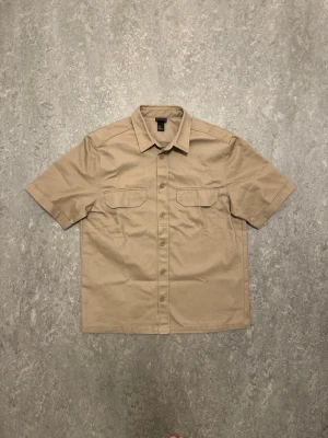Beige kortärmad skjorta med fickor - Stilren beige skjorta från H&M med korta ärmar och två bröstfickor med lock. Skjortan har klassisk krage och knäppning framtill. Perfekt för en avslappnad och trendig look.