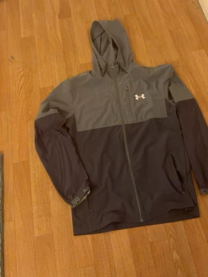Grå och svart vindjacka Under Armour - Snygg vindjacka från Under Armour i tvåfärgad design med grå upptill och svart nedtill. Jackan har huva, hel dragkedja framtill och två sidofickor. Under Armour-logga på bröstet och diskreta detaljer på ärmen. Perfekt för blåsiga dagar.