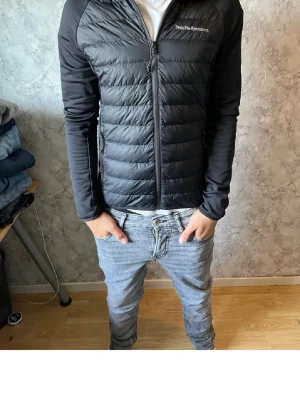 Peak performance cardigan jacka - Snygg svart pufferjacka från Peak Performance med quiltad front och rygg samt släta ärmar. Jackan har dragkedja framtill, två sidofickor med dragkedja och en diskret logga på bröstet. Perfekt för kalla dagar och enkel att matcha med olika outfits.