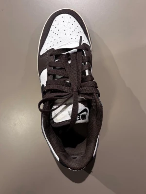 Nike Dunk Low mörkbrun/vit sneaker - Snygga Nike Dunk Low sneakers i mörk brun och vitt med klassisk Swoosh på sidan. Ovandelen är i läder med perforerad tåbox för extra ventilation. Skon har svart yttersula och matchande svarta skosnören. Perfekt för dig som gillar streetwear och tidlös design.