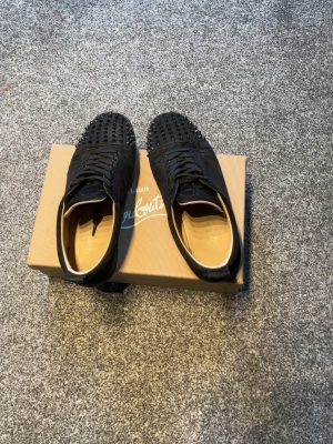 Svarta Louboutin sneakers med nitar - Säljer ett par svarta sneakers från Christian Louboutin med coola svarta nitar över hela tån och ovandelen. Skorna har snörning och är gjorda i mocka med klassisk röd sula. Originalbox kommer med.
