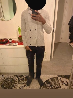 Kofta - Säljer en stilren ljusgrå kofta med dubbelknäppning och svarta knappar. Koftan har ribbade muddar vid ärmslut och nederkant samt två sidofickor. Perfekt för lager på lager och enkel att matcha med olika outfits.