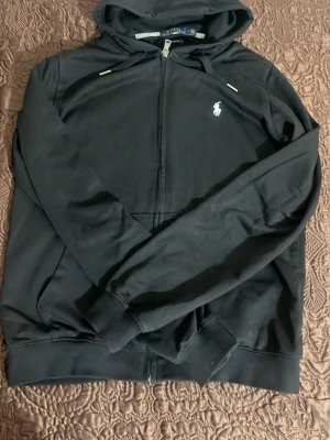 Svart hoodie från Polo Ralph Lauren - Svart hoodie med dragkedja från Polo Ralph Lauren. Klassisk design med huva och dragsnören, samt vit broderad logga på bröstet. Tillverkad i mjukt material och har ribbade muddar vid ärmslut och nederkant. Perfekt för en avslappnad och stilren look.