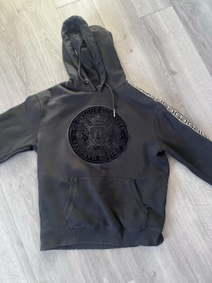 Svart hoodie Glorious Gangsta - Svart hoodie från Glorious Gangsta med stor, broderad logga i sammet på bröstet och mönstrade band längs ärmarna. Klassisk känguruficka framtill och justerbar huva med dragsko. Materialet är mjukt och bekvämt, perfekt för en streetwear-look.
