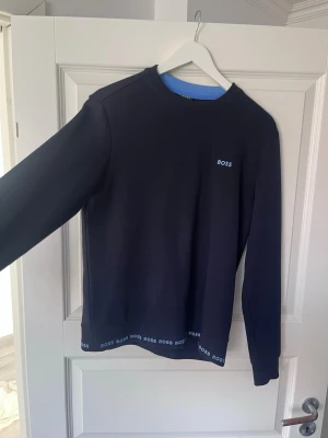Svart sweatshirt från BOSS - Stilren svart sweatshirt från BOSS med rund halsringning och diskret logga på bröstet. BOSS-tryck i blått längs nederkanten och halsen. Perfekt för en clean och modern look. Mjukt material och lång ärm.