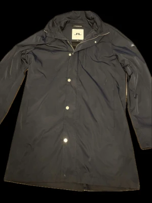 Mörkblå jacka från J.Lindeberg - Stilren mörkblå jacka från J.Lindeberg med huva och tryckknappar framtill. Jackan har diskret logga på ärmen och är tillverkad i vattenavvisande material. Perfekt för regniga dagar när du vill hålla dig torr och snygg.