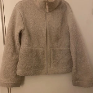 Beige teddyjacka med dragkedja - Mysig beige teddyjacka med hög krage och hel dragkedja framtill. Jackan har två stora fickor på framsidan och är tillverkad i fluffigt teddy-material. Perfekt för kyliga dagar och ger en avslappnad vibe.