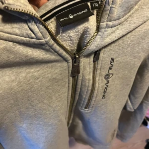 Grå hoodie från Sail Racing - Snygg grå hoodie från Sail Racing med dragkedja framtill och bröstficka med zip. Tröjan har huva, långa ärmar och logga på bröstet samt på huvan. Perfekt för chill eller när du vill ha en enkel och stilren look.