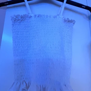 Vit smockad linnetopp med volang - Supersöt vit linnetopp från XOXO Friends med smockad resår över hela toppen och volangkant nertill. Axelbanden är smala och toppen har en luftig känsla, perfekt för varma dagar. Enkel att matcha med jeans eller kjol.