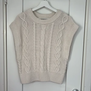 Stickad beige väst från & Other Stories - Säljer en chunky stickad väst från & Other Stories i beige. Västen har bred ribbad krage, ärmlösa kanter och snyggt flätstickat mönster framtill. Perfekt att slänga över en skjorta eller t-shirt för en chill och trendig look. Knappt använd.