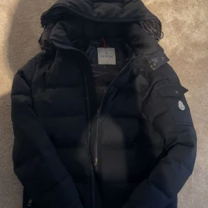 Svart Moncler pufferjacka med huva - Säljer en svart pufferjacka från Moncler med huva och dragkedja. Jackan har Moncler-logga på ärmen, praktisk ficka med knapp på ärmen och quiltad design. Perfekt för kalla dagar och riktigt snygg streetstil.