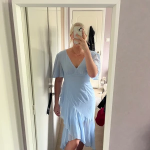 Ljusblå klänning  - Säljer en ljusblå klänning med omlott-design och v-ringning. Klänningen har korta, fladdriga ärmar och volangdetaljer längs nederdelen. Materialet är tunt och ger ett luftigt intryck. Använt 1 gång på bröllop. Med dragkejda i ryggen. Tveka inte att höra av dig med frågor.☺️