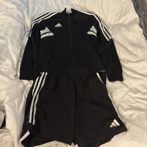 Svarta Adidas träningset - Svarta träningsshorts från Adidas med klassiska vita ränder längs sidorna och logga på benet. Shortsen har elastisk midja med snörning och är gjorda i lätt polyester som andas. Perfekta för sport eller gym. Snygg och sportig look.